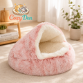 A pink plush pet bed with a cream-colored interior, styled in a warm, sunlit room with a wooden side table, lamp, and potted plant. The CozyDen brand logo is visible in the upper left corner.